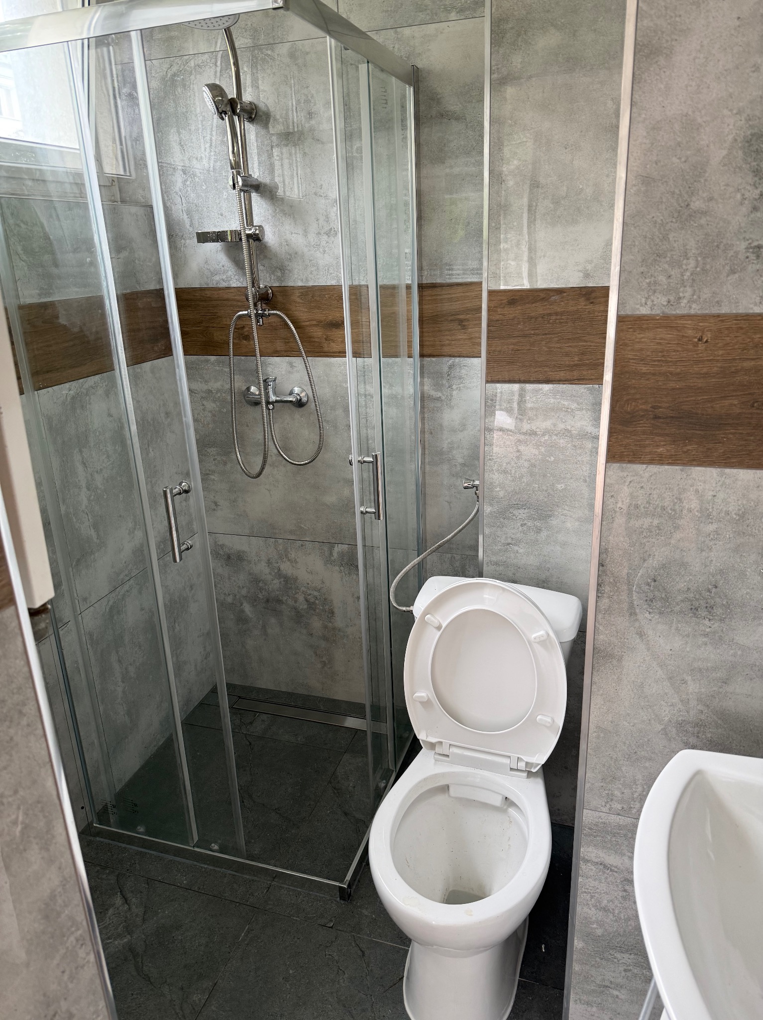 Nowoczesna łazienka po remoncie: kabina prysznicowa ze szklanymi drzwiami, toaleta z otwartą klapą i umywalka. Szare płytki z drewnianym akcentem na ścianach.