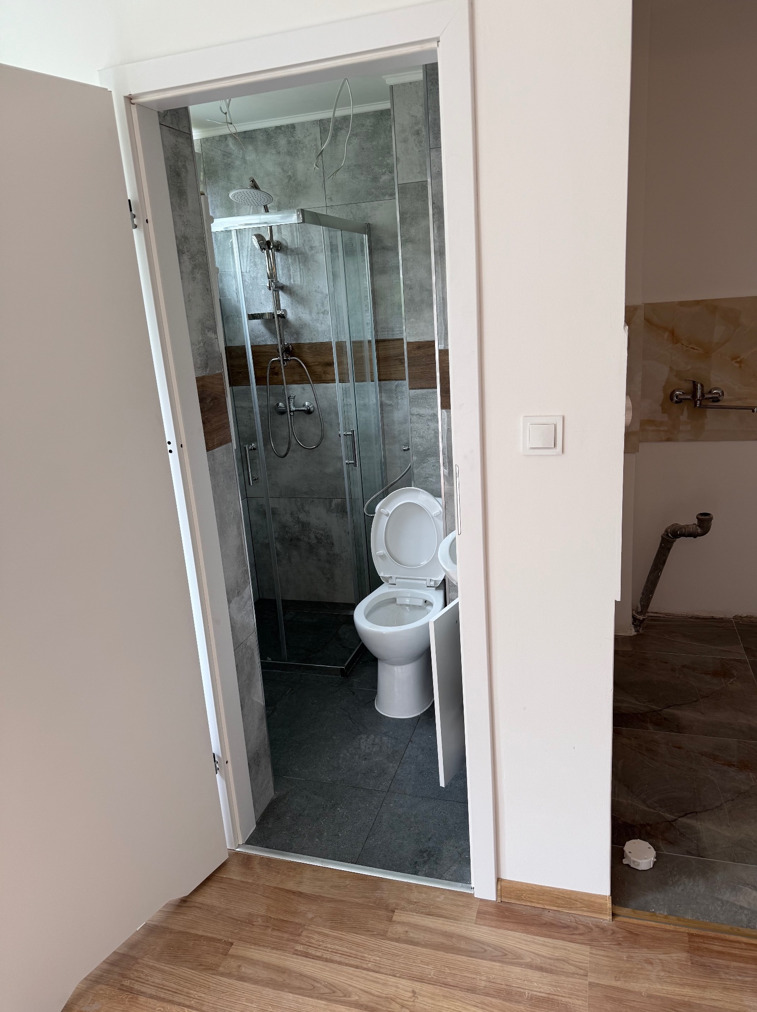 Otwarta łazienka z kabiną prysznicową i toaletą w odcieniach szarości i brązu, widoczna z korytarza z panelami podłogowymi i niedokończoną ścianą z instalacją.