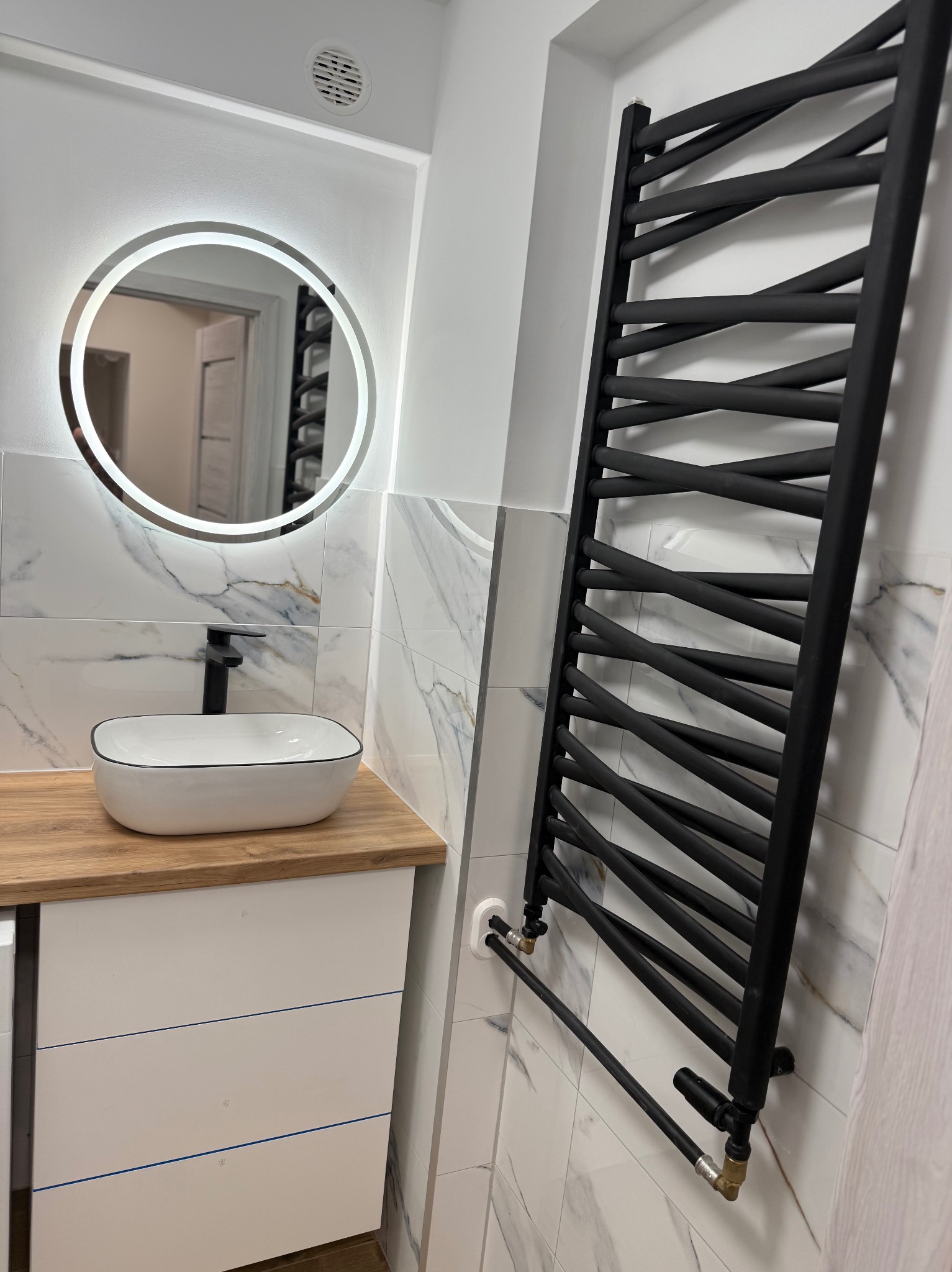 Nowoczesna łazienka po remoncie: biała umywalka nablatowa, lustro z podświetleniem LED i czarny, designerski grzejnik na tle marmurowych płytek.