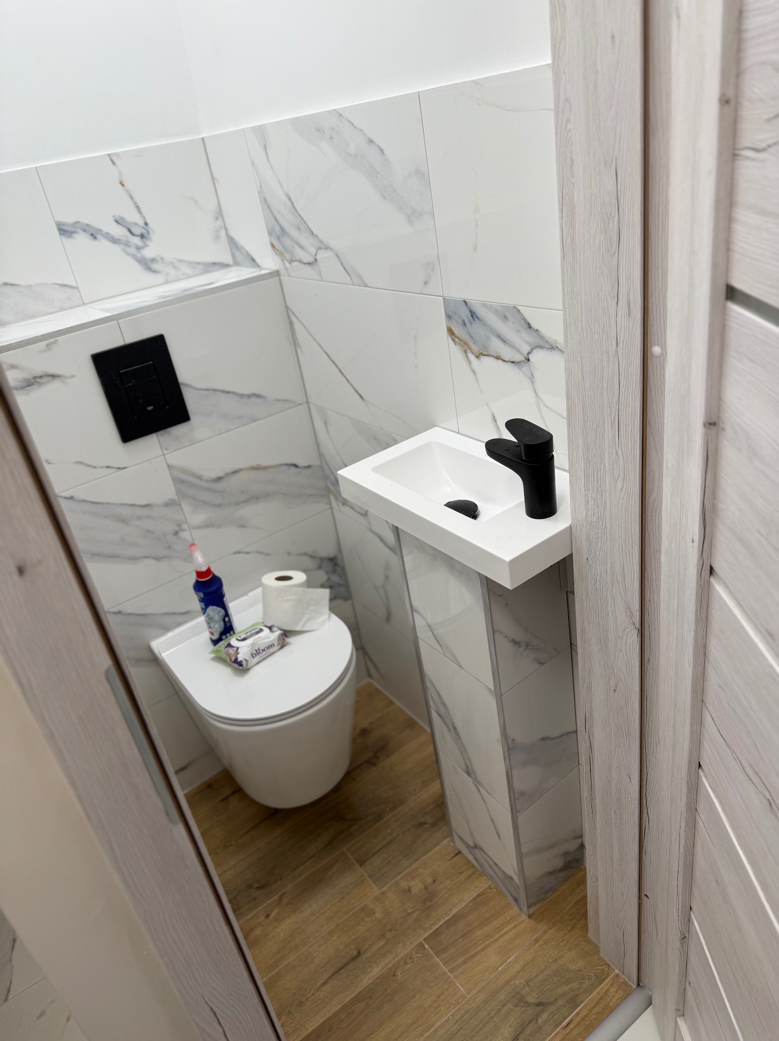 Nowoczesna, wykończona łazienka z umywalką na postumencie i marmurowymi płytkami. Uwagę przykuwa czarna armatura i drewnopodobne panele podłogowe.