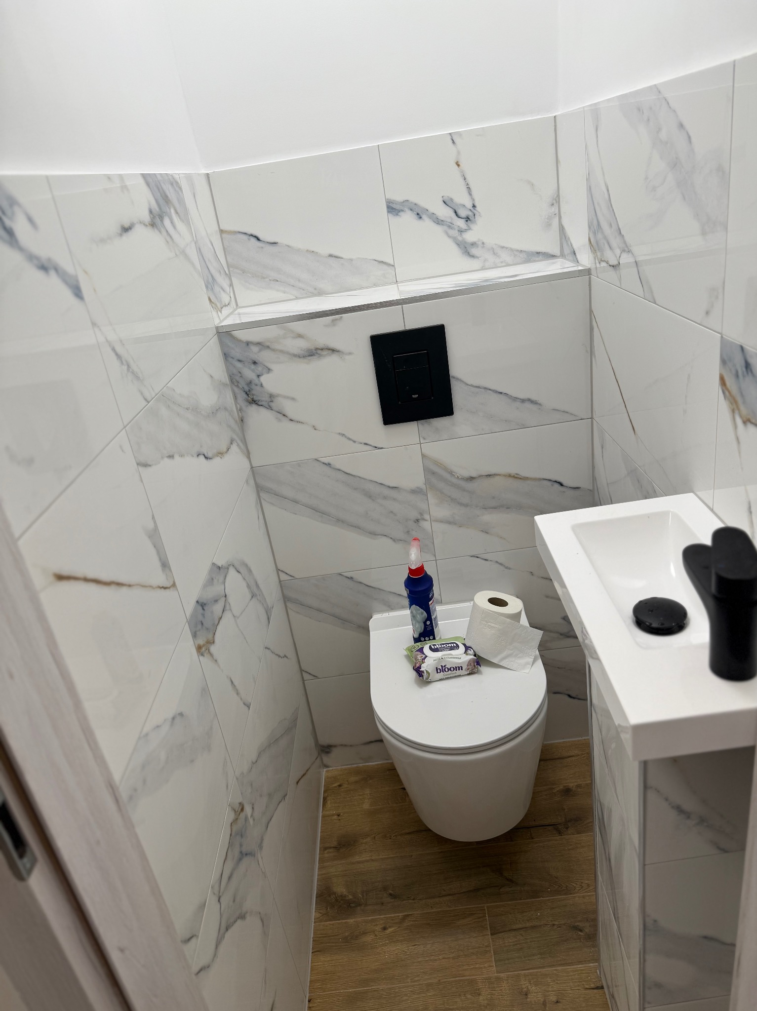 Wykończona, mała toaleta z marmurowymi płytkami na ścianach, drewnopodobną podłogą, wiszącą miską WC i umywalką z czarną armaturą. Nowoczesny design, wąskie pomieszczenie.
