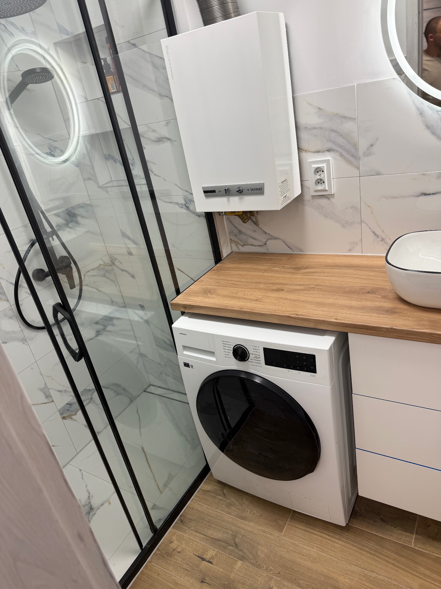 Nowoczesna łazienka z kabiną prysznicową typu walk-in, pralką pod blatem z drewna i białymi płytkami imitującymi marmur. Oświetlone lustro i piec gazowy.