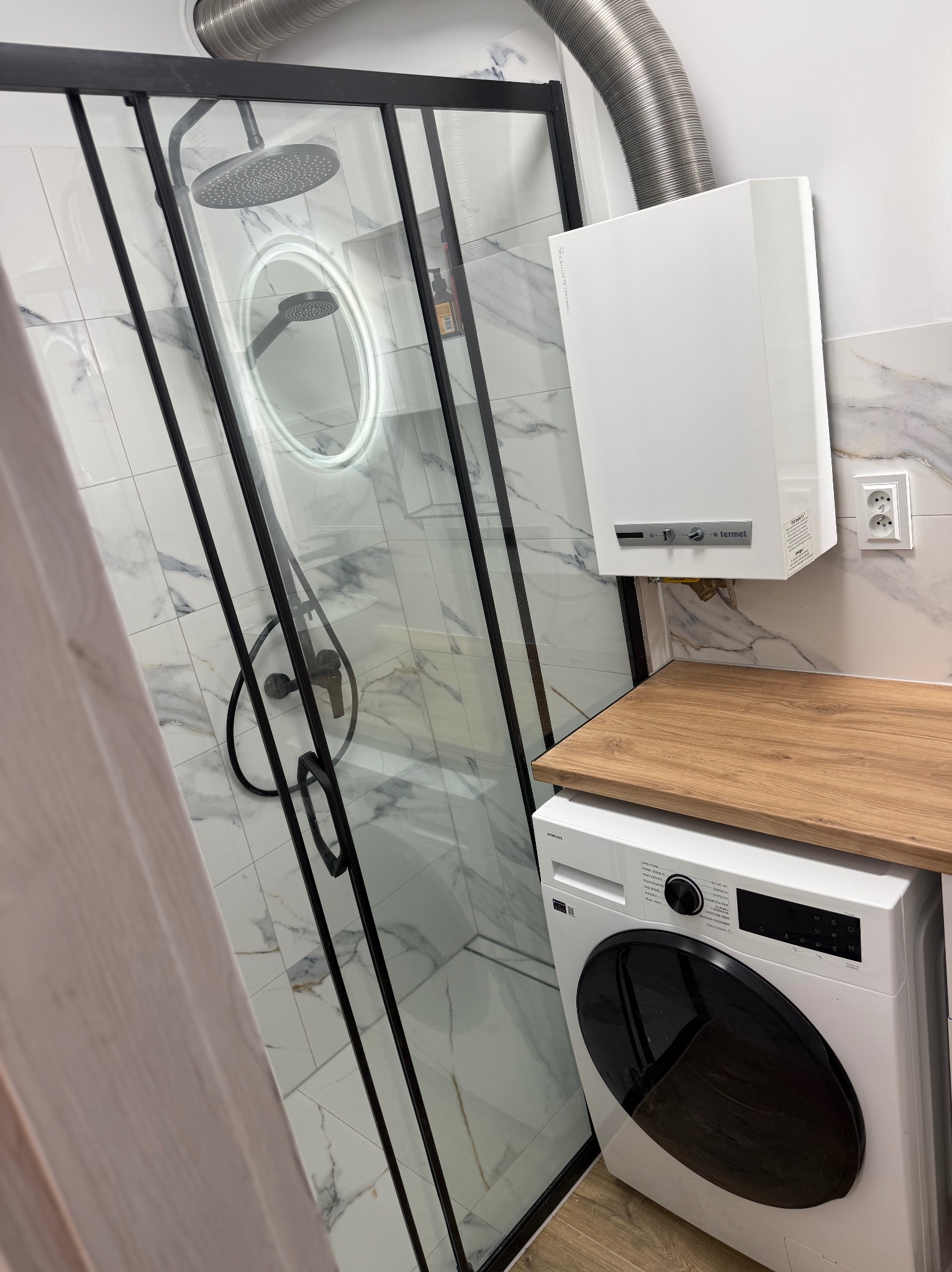 Nowoczesna łazienka z kabiną prysznicową typu walk-in z czarnymi ramami, marmurowymi płytkami i pralką pod drewnianym blatem. Widoczny bojler i oświetlenie LED.