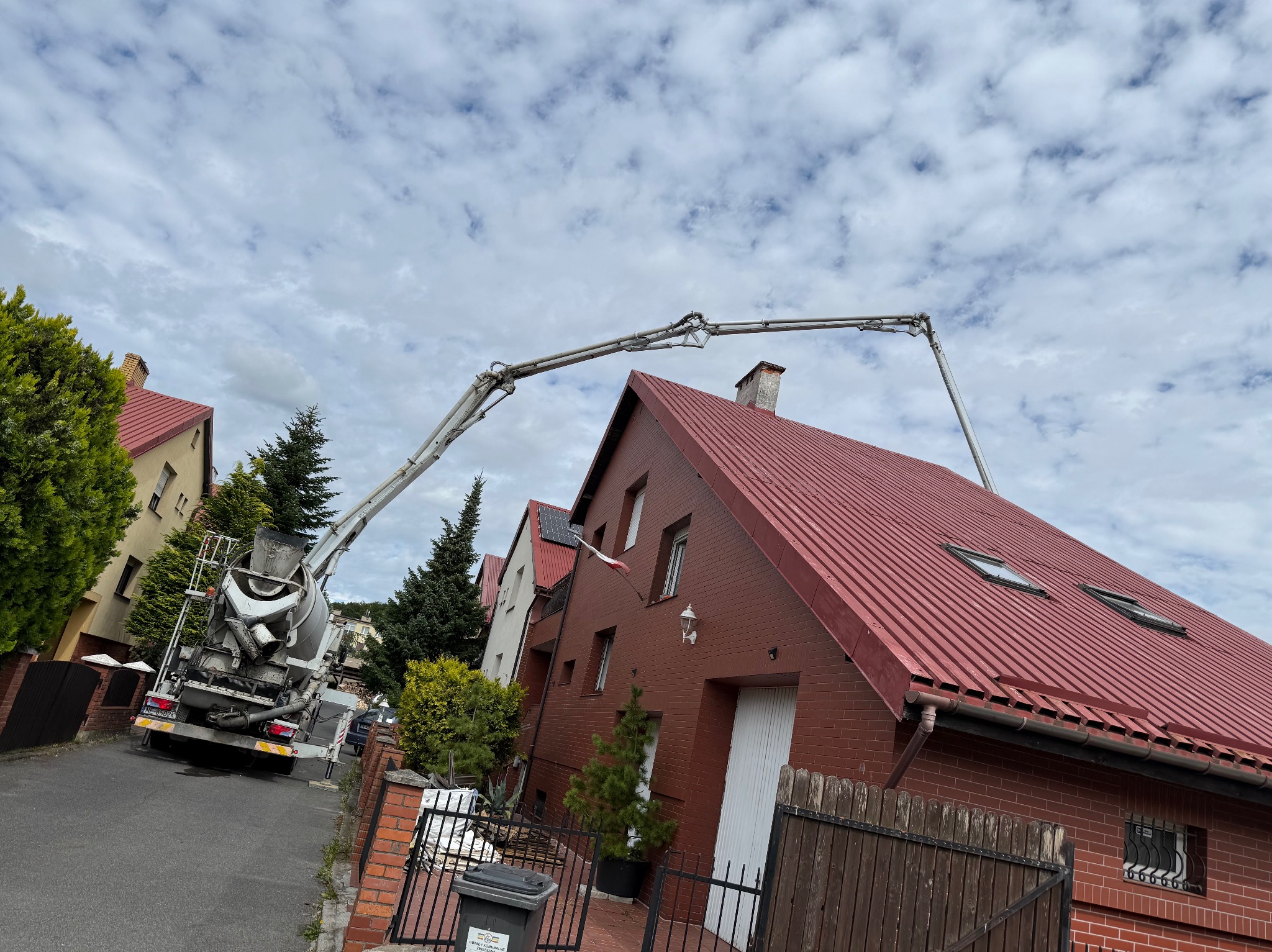Betonomieszarka z wysięgnikiem pompuje beton na dach domu z czerwoną blachodachówką i oknami dachowymi, widok z ulicy w pochmurny dzień.