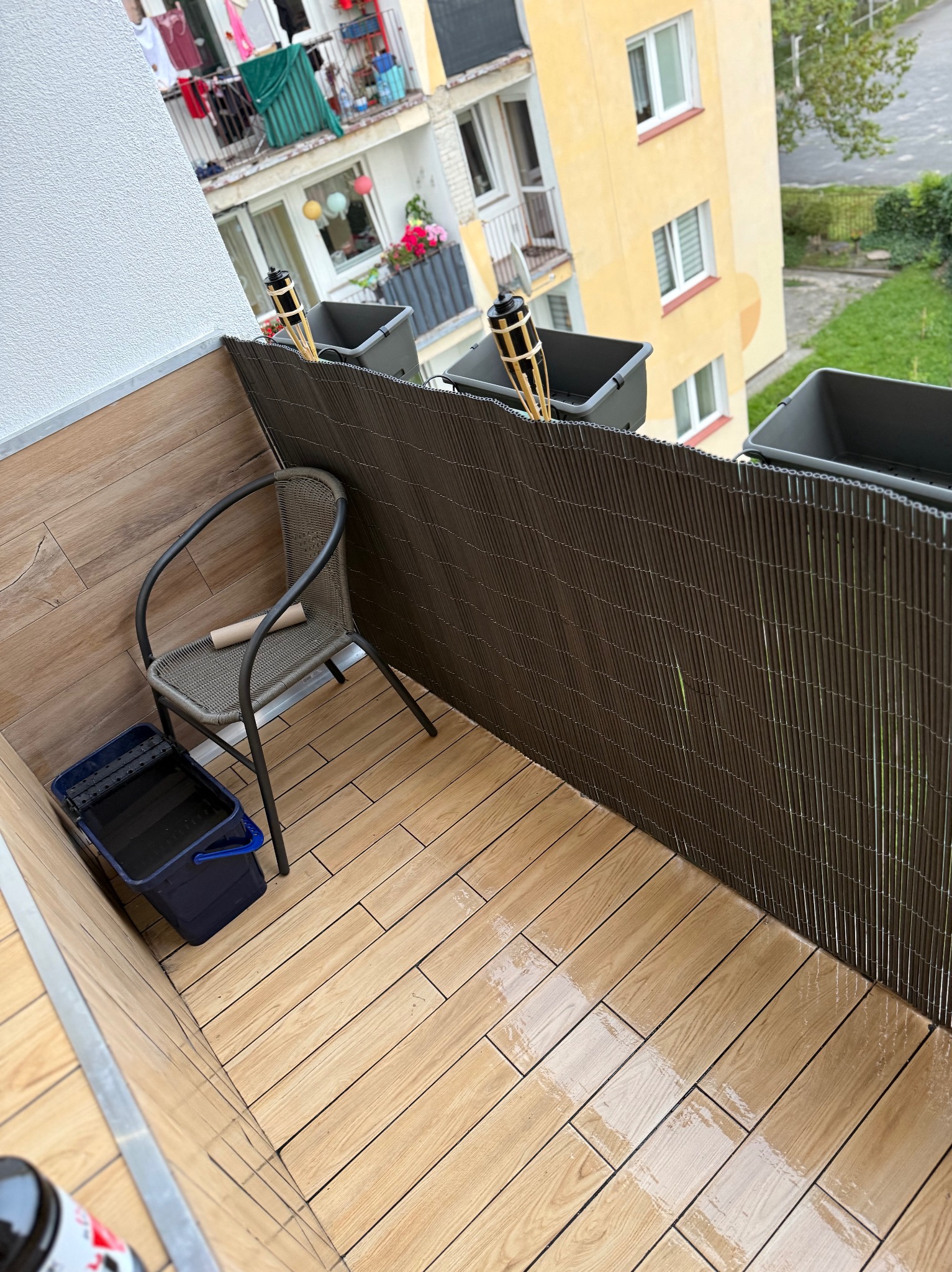 Wykończony balkon z drewnopodobnymi płytkami, brązową osłoną i krzesłem. Widoczny kubeł z akcesoriami malarskimi. Dekoracyjne lampiony w donicach.