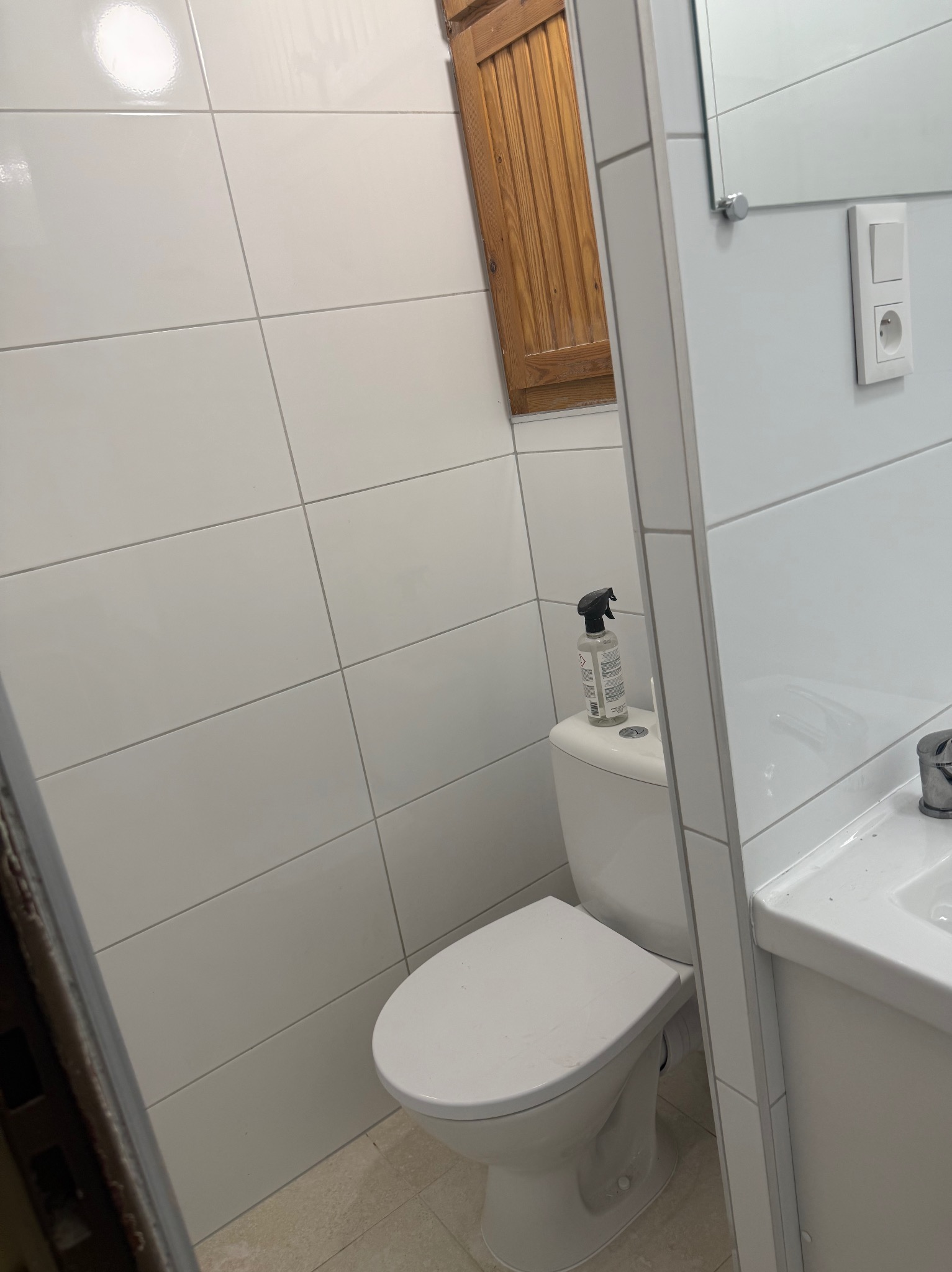 Biała toaleta z deską sedesową i dozownikiem płynu na spłuczce, na tle ściany z białych płytek, w odnowionej łazience z drewnianą szafką w tle.