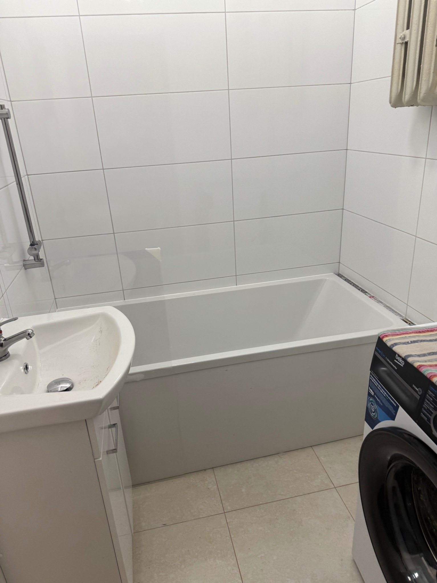 Biała łazienka po remoncie: wanna, umywalka z szafką, pralka Beko. Białe płytki na ścianach i jasne płytki na podłodze. Grzejnik na ścianie.