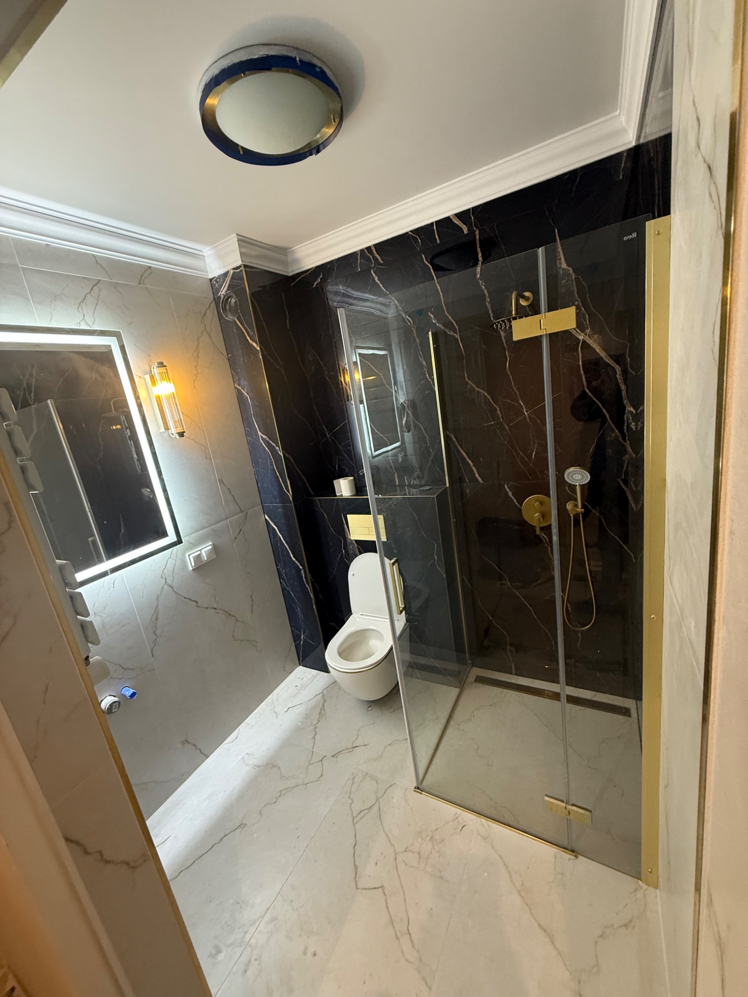 Elegancka łazienka z kabiną prysznicową w złotej ramie, marmurowymi płytkami i podświetlanym lustrem. Widoczna toaleta i nowoczesne oświetlenie sufitowe.