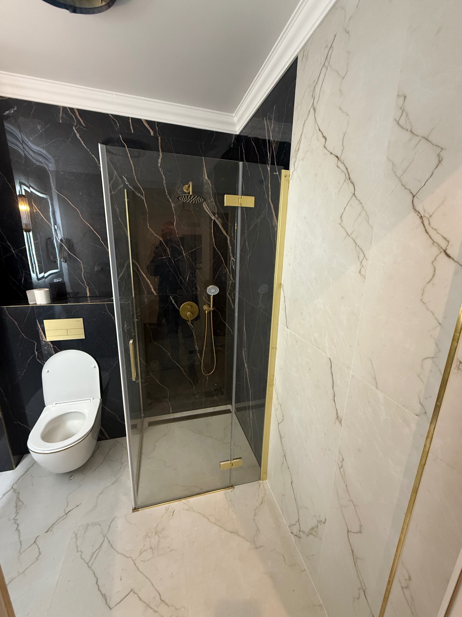 Elegancka łazienka z kabiną prysznicową typu walk-in z złotymi detalami, marmurowymi płytkami w odcieniach bieli i czerni oraz podwieszaną toaletą.