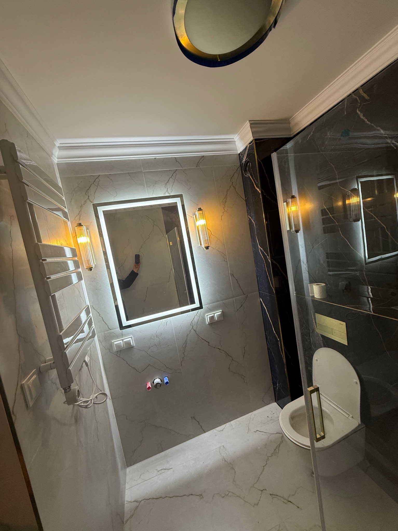 Elegancka łazienka z marmurowymi płytkami, podświetlanym lustrem i grzejnikiem drabinkowym. Widoczne detale wykończenia i oświetlenie.