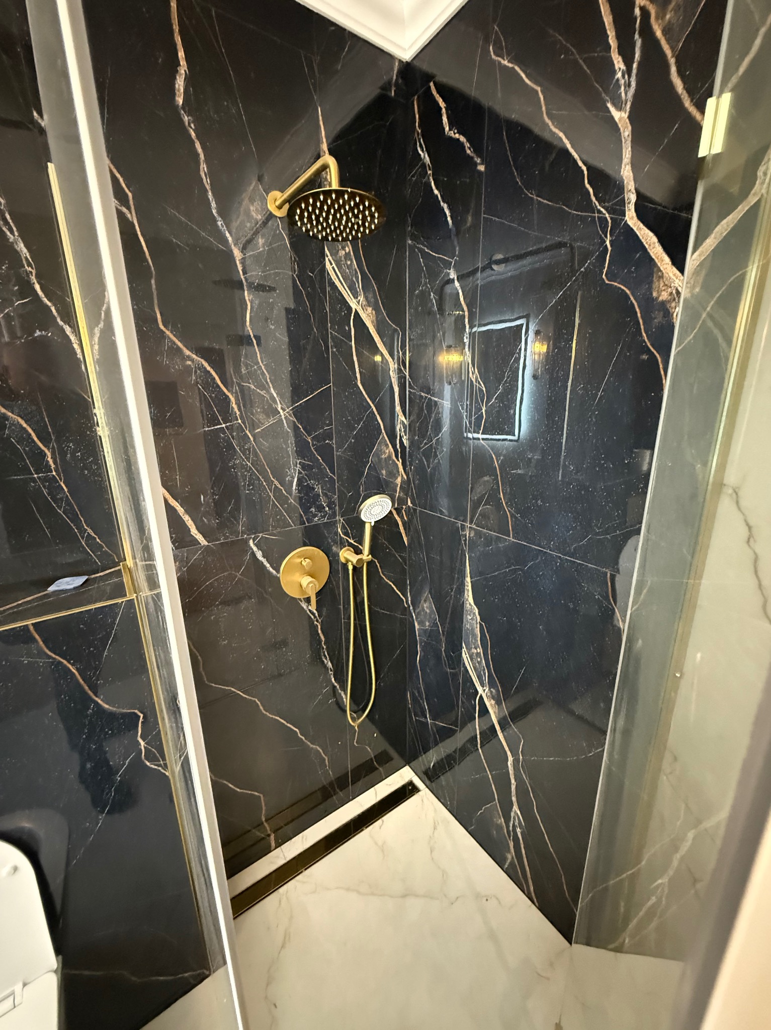Kabina prysznicowa z czarnymi płytkami imitującymi marmur z złotymi żyłami i złotą armaturą. Widok z góry, podkreślający luksusowy charakter wykończenia.