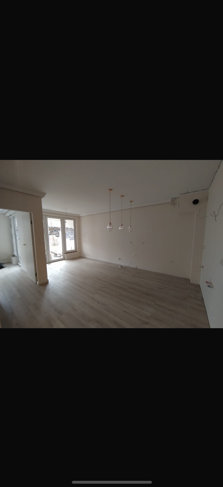 Jasne panele podłogowe w przestronnym, odświeżonym pokoju z trzema wiszącymi lampami. Świeżo pomalowane ściany i duże okno balkonowe w tle. Widoczne ślady po instalacji elektrycznej.