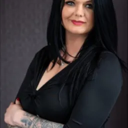 Portret kobiety z ciemnymi włosami i tatuażami na ramionach, ubrana w czarną bluzkę i spódnicę w kropki, pozująca z założonymi rękami na tle szarej ściany.