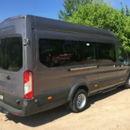 Szary bus Ford Transit z przyciemnianymi szybami, widziany z tyłu pod kątem, na piaszczystym podłożu, z budynkiem w tle.