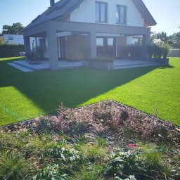 GARDEN LAWN & IRRIGATION MARCIN PIECHOTA - Nowoczesny ogród z równo przystrzyżonym trawnikiem, rabatami z korą i różnorodną roślinnością oraz domem w tle.