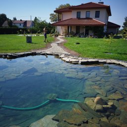 GARDEN LAWN & IRRIGATION MARCIN PIECHOTA - Oczko wodne z kamieniami na dnie i zielonym wężem, w tle trawnik, ścieżka z kamienia prowadząca do domu i pracownik z wężem w ręku.