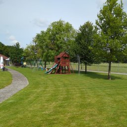 GARDEN LAWN & IRRIGATION MARCIN PIECHOTA - Rozległy, zadbany trawnik z automatycznym systemem nawadniania, plac zabaw z drewnianą wieżą i zjeżdżalnią, oraz ścieżka z kostki brukowej prowadząca do altany z czerwonym dachem.