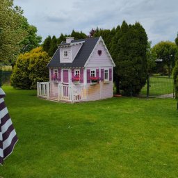 GARDEN LAWN & IRRIGATION MARCIN PIECHOTA - Różowy domek dla dzieci z ciemnym dachem i kwiatami w oknach, otoczony zielonym trawnikiem i drzewami.