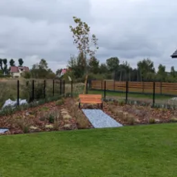 Nowoczesny ogród z drewnianą ławką, ścieżką z szarego żwiru, młodym drzewkiem podpartym palikiem, rabatami wysypanymi korą, robotem koszącym trawę i ogrodzeniem panelowym.