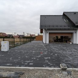 GARDEN LAWN & IRRIGATION MARCIN PIECHOTA - Nowa, szara kostka brukowa ułożona na podjeździe przed domem z otwartym garażem, widoczne palety w tle i skrzynka elektryczna przy ogrodzeniu.