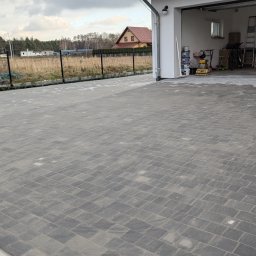 GARDEN LAWN & IRRIGATION MARCIN PIECHOTA - Nowo ułożona, szara kostka brukowa przed garażem z widocznymi śladami piasku i niewielkim fragmentem gruntu z kamieniami przy krawędzi.