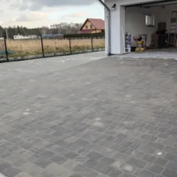 Nowo ułożona, szara kostka brukowa przed garażem z widocznymi śladami piasku i niewielkim fragmentem gruntu z kamieniami przy krawędzi.