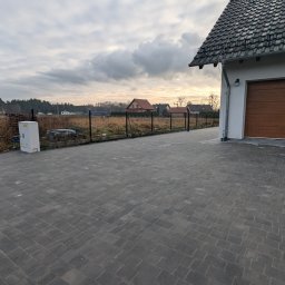 GARDEN LAWN & IRRIGATION MARCIN PIECHOTA - Ułożona kostka brukowa w kolorze grafitowym na podjeździe, widoczny fragment domu z brązową bramą garażową, ogrodzenie panelowe w tle i zachmurzone niebo.