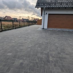 GARDEN LAWN & IRRIGATION MARCIN PIECHOTA - Ułożona szara kostka brukowa na podjeździe w Dobrzeniu Wielkim, widoczny fragment domu z brązową bramą garażową i ogrodzenie panelowe w tle.