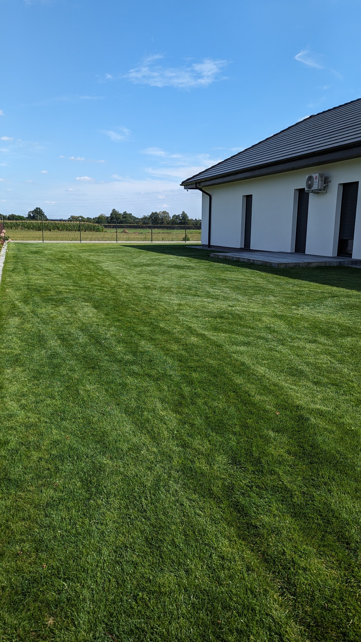 Perfekcyjnie przystrzyżony trawnik z wyraźnymi pasami, przylegający do nowoczesnego domu z białymi ścianami i ciemną dachówką, w tle widoczne pole i błękitne niebo.
