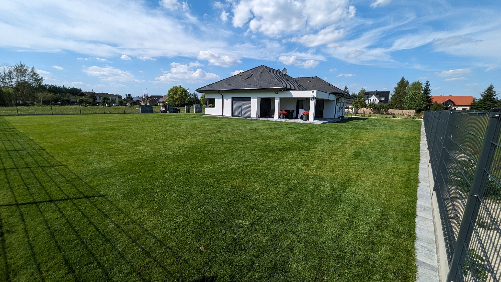 Rozległy trawnik z rolki przed nowoczesnym domem jednorodzinnym z ogrodzeniem panelowym w słoneczny dzień.