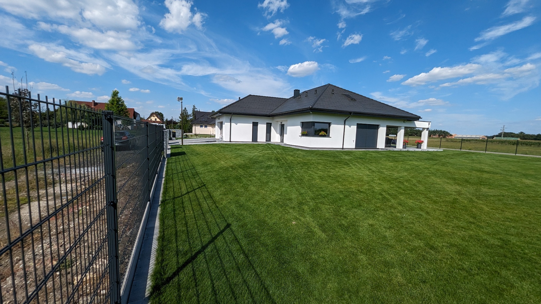 Nowoczesny dom jednorodzinny z zadbanym trawnikiem i ogrodzeniem panelowym w słoneczny dzień.