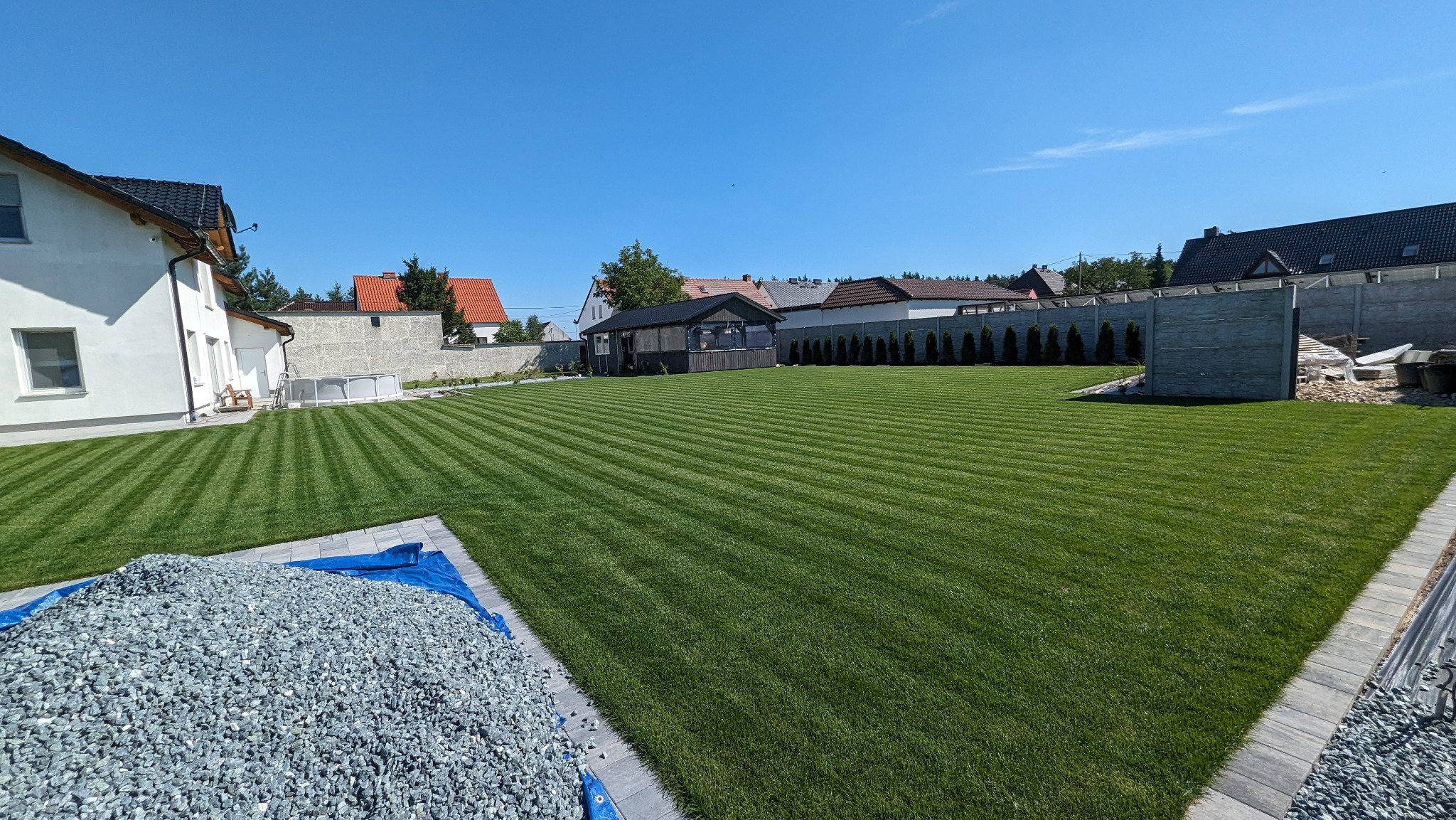 Perfekcyjnie przystrzyżony trawnik z wyraźnymi pasami, z widocznym stosem grysu na pierwszym planie i altaną w tle, otoczony ogrodzeniem panelowym i tujami.