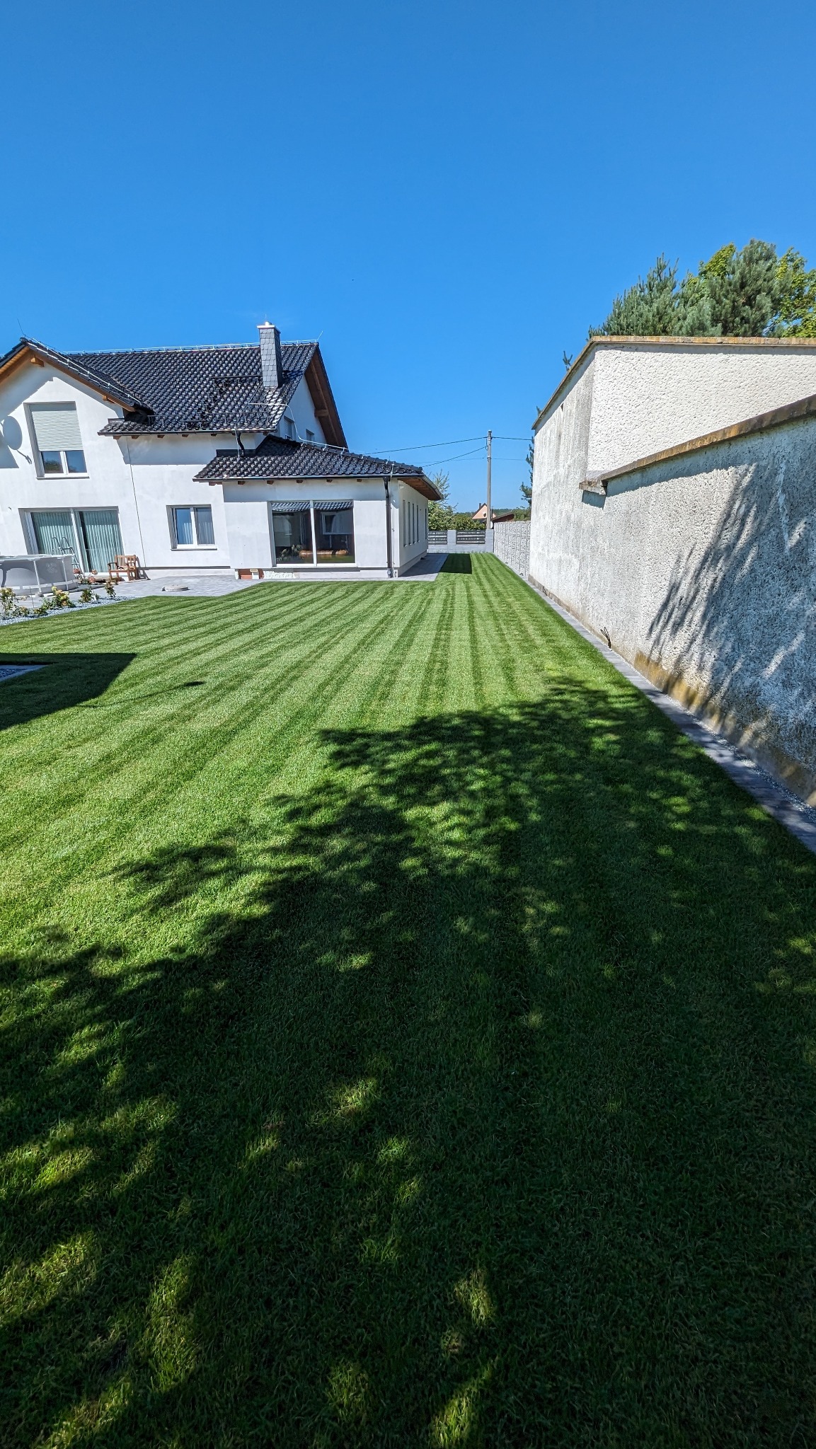 Perfekcyjnie przystrzyżony trawnik w paski przed nowoczesnym białym domem z ciemnym dachem, widok z cienia drzewa.