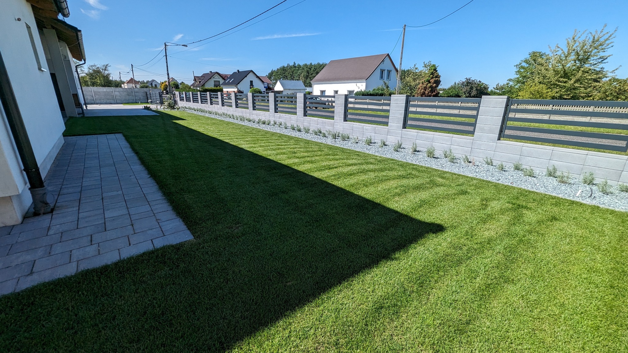 Nowoczesny ogród z równo przystrzyżonym trawnikiem, szarym ogrodzeniem panelowym na betonowej podmurówce, ozdobnym żwirem i nasadzeniami, w tle budynki mieszkalne.