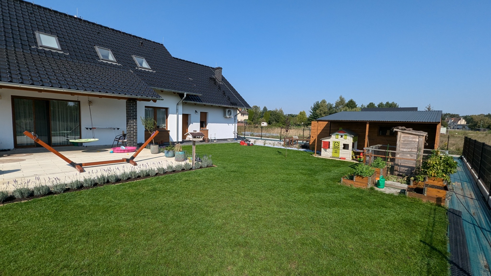 Starannie utrzymany trawnik przed nowoczesnym domem z ciemną dachówką, widoczny drewniany hamak, rabaty z lawendą, domek dla dzieci i drewniana szopa na narzędzia w głębi ogrodu.