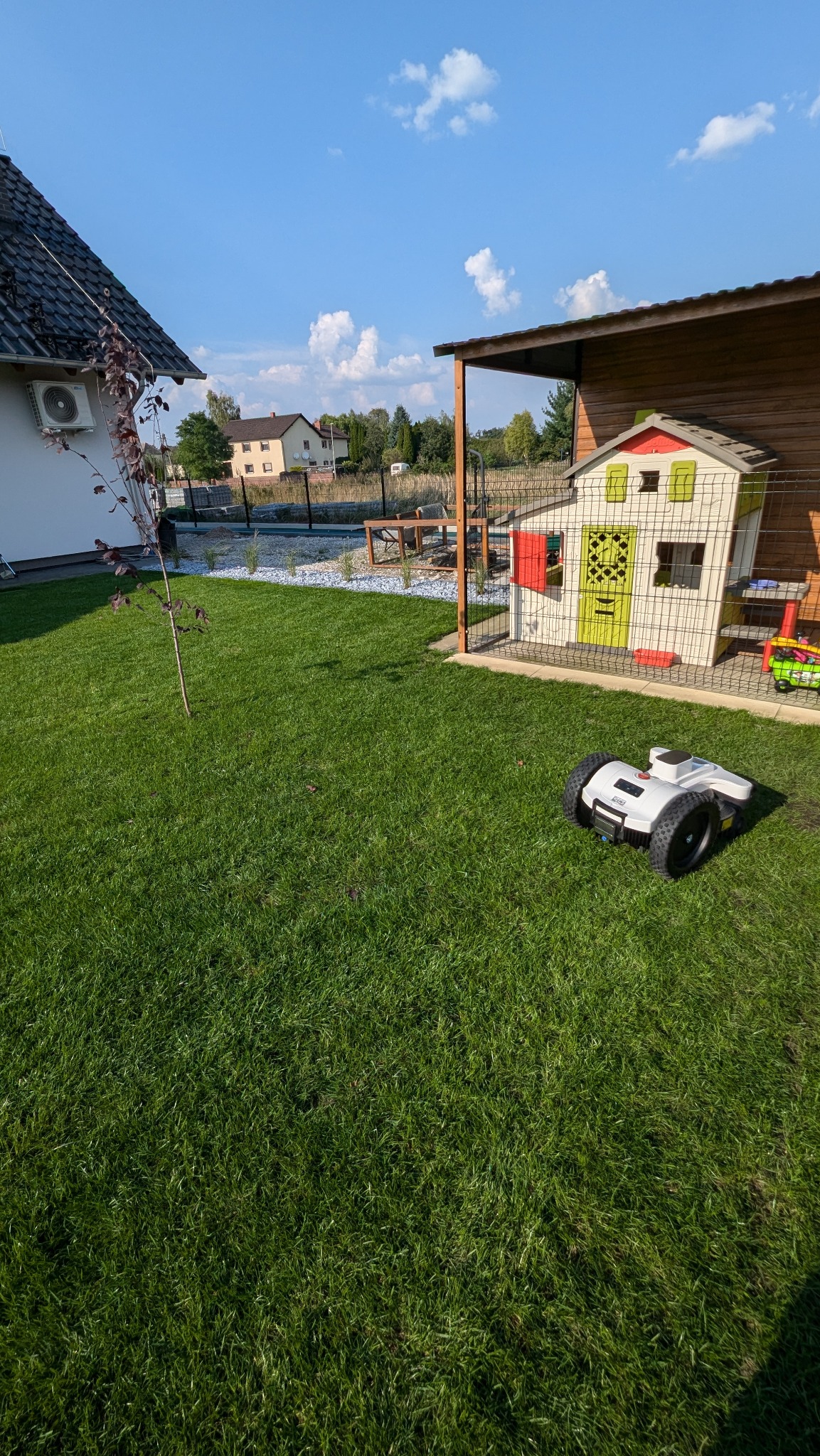 Robot koszący na zadbanym trawniku, w tle domek dla dzieci, nasadzenia z białym kamieniem i budynki mieszkalne w słoneczny dzień.
