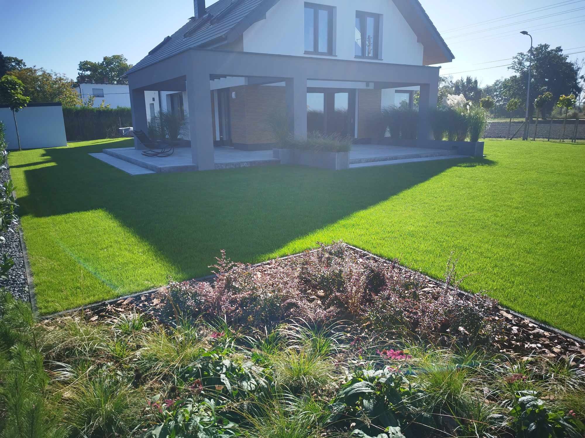 Nowoczesny ogród z równo przystrzyżonym trawnikiem, rabatami z korą i różnorodną roślinnością oraz domem w tle.