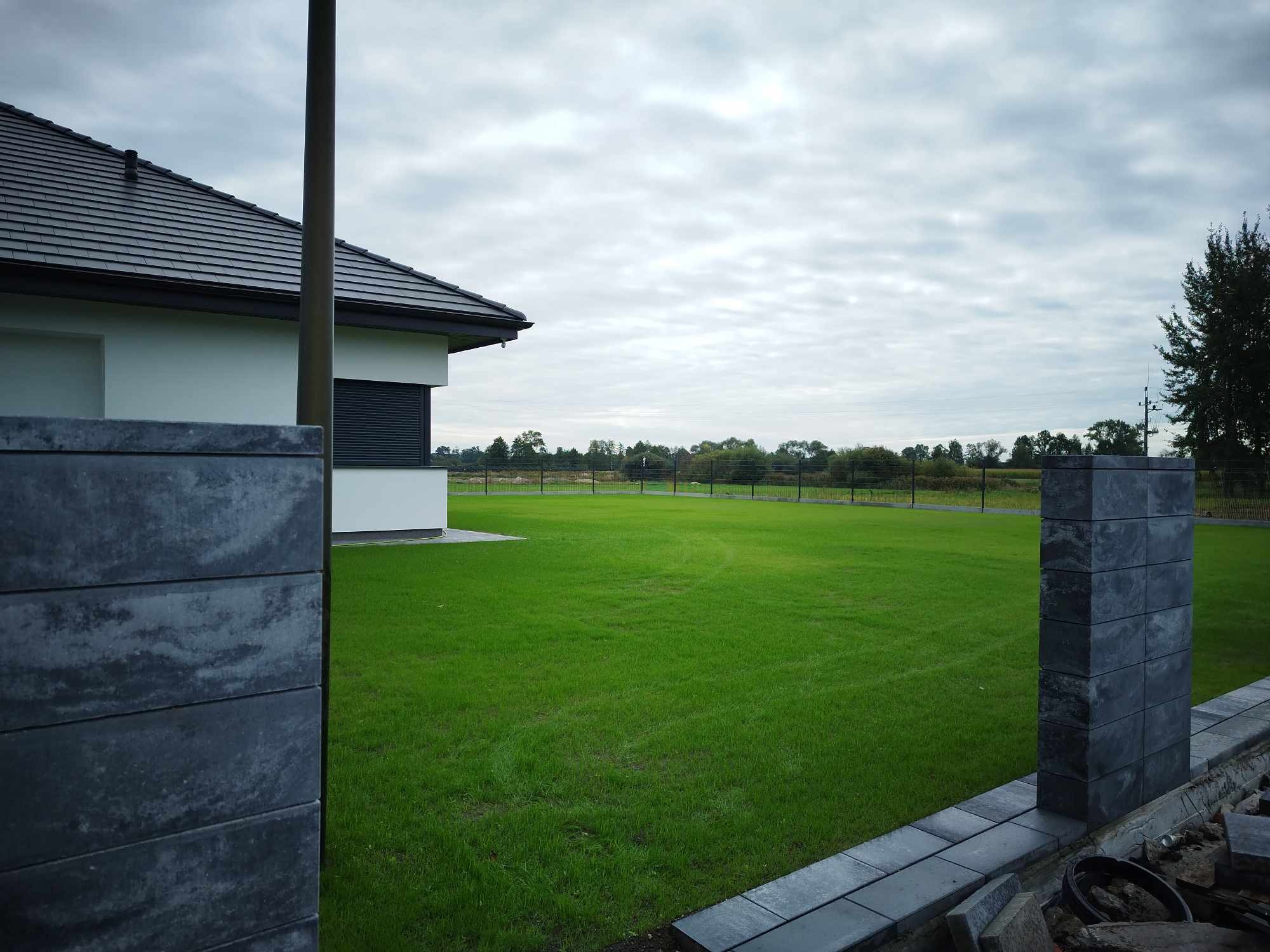 Widok na świeżo założony trawnik z rolki, otoczony nowoczesnym ogrodzeniem z paneli betonowych i domu z ciemną dachówką, w tle widoczne drzewa.