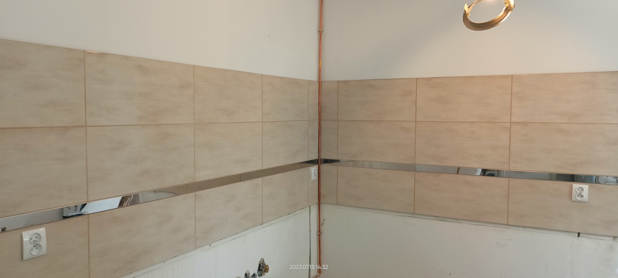 Jasne, beżowe płytki ścienne z błyszczącą, metalową listwą dekoracyjną, widoczne gniazdka elektryczne i miedziana rurka przy ścianie, w dolnej części ściany widoczne ślady po remoncie.