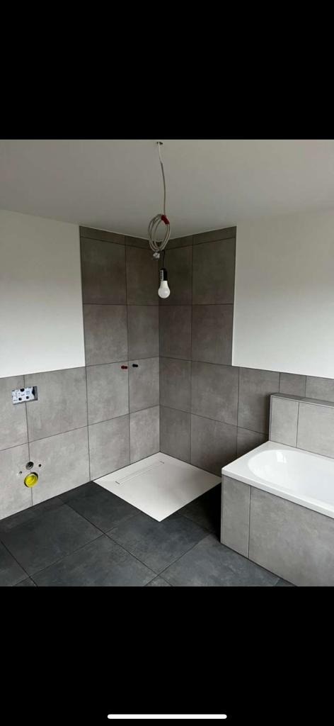 Szare płytki ścienne i ciemne podłogowe w łazience w trakcie remontu, widoczne instalacje wodne i elektryczne, surowa żarówka zwisająca z sufitu, wanna obudowana płytkami, biały brodzik.