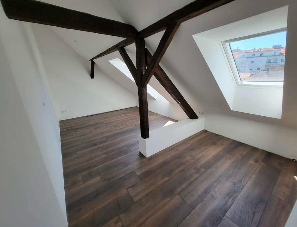 Poddasze po remoncie: brązowe panele podłogowe ułożone w jodełkę, białe ściany, drewniana konstrukcja stropu, okno dachowe z widokiem na budynki.