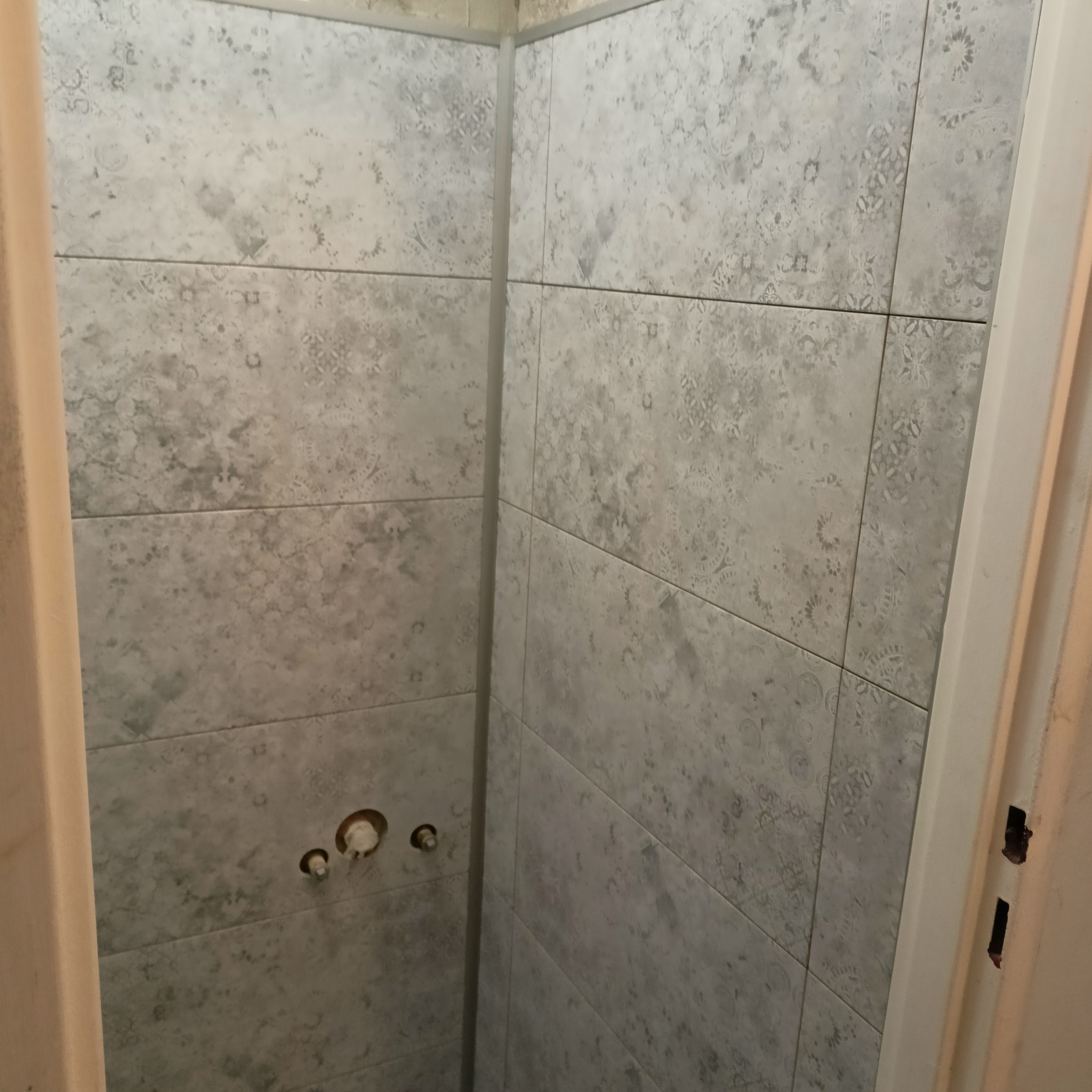 Ściana łazienki wyłożona szarymi płytkami z delikatnym wzorem, widoczne otwory na baterię, ujęcie z bliska.