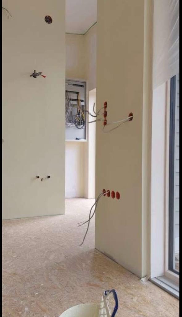 Wnętrze w trakcie prac wykończeniowych: ściany z otworami na instalacje elektryczne, widoczne przewody, surowa podłoga, skrzynka z bezpiecznikami w tle.