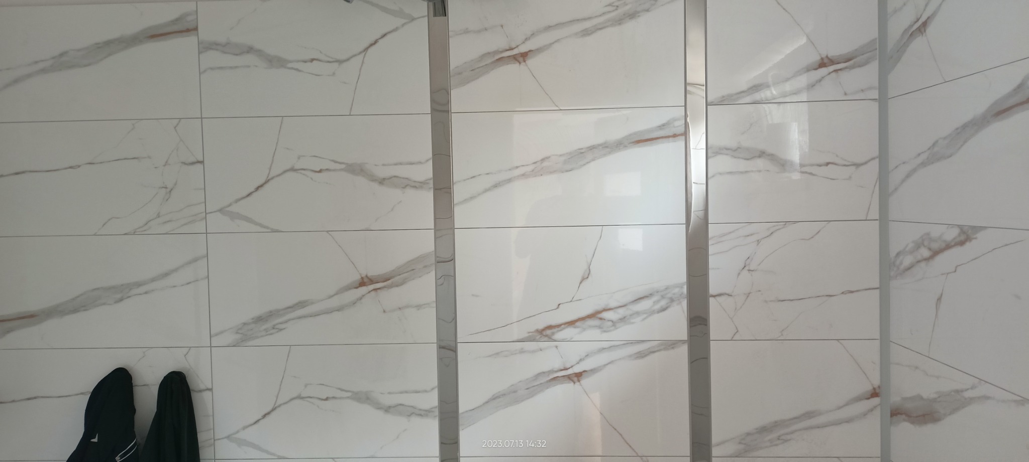 Położone białe płytki imitujące marmur z szarymi i rdzawymi żyłkami, oddzielone srebrnymi listwami, z fragmentem czarnej kurtki w lewym dolnym rogu.