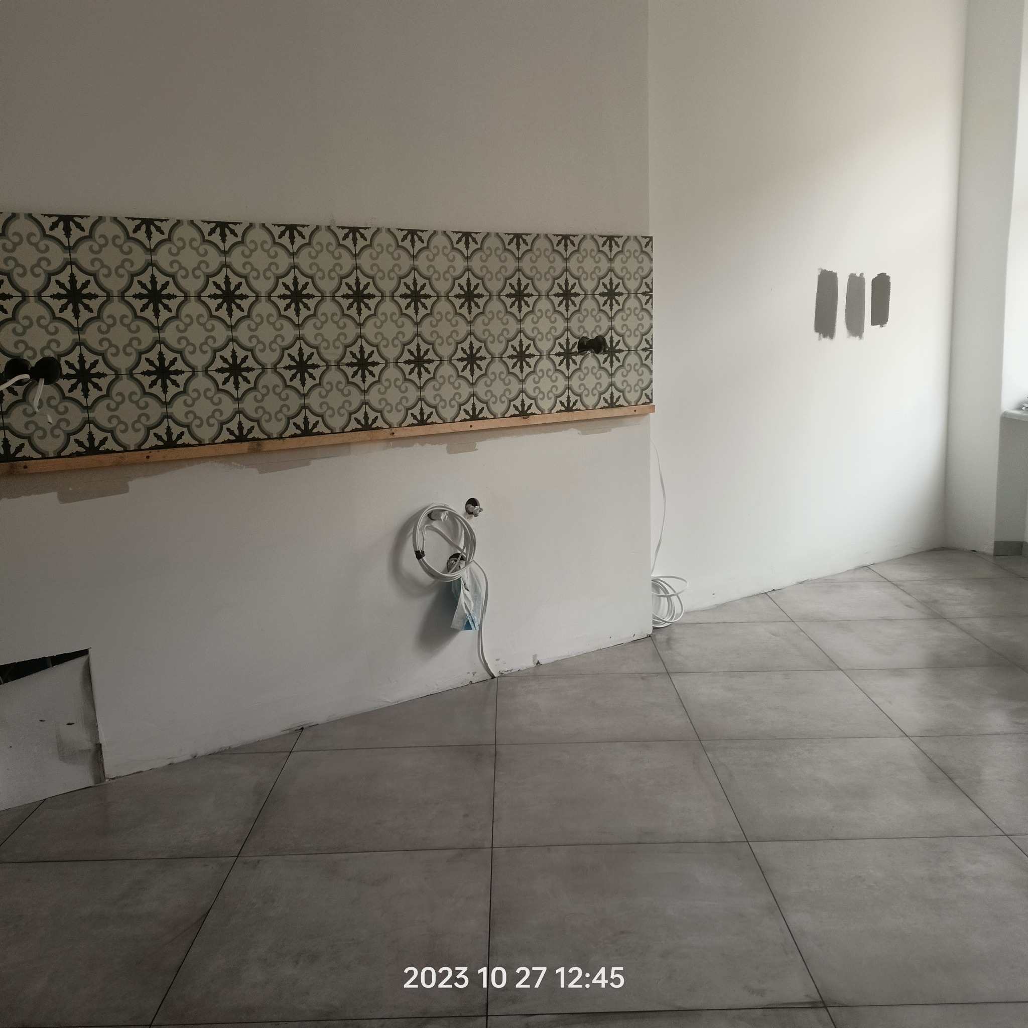 Fragment kuchni w trakcie prac wykończeniowych: położone szare płytki podłogowe, fragment ściany z dekoracyjnymi płytkami ściennymi z motywem roślinnym, widoczne przygotowane instalacje...