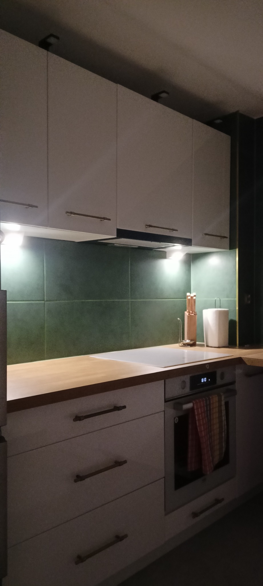 Nowoczesna kuchnia z białymi szafkami, drewnianym blatem i zielonymi płytkami, oświetlona punktowymi lampami podszafkowymi, z widocznymi akcesoriami kuchennymi i piekarnikiem z ręcznikiem.