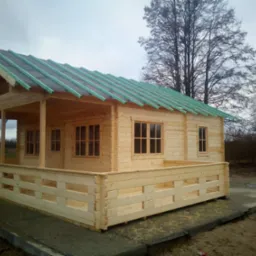 Surowy drewniany domek letniskowy z niezakończonym dachem pokrytym papą i listwami, otoczony drewnianą balustradą, na betonowej wylewce, z drzewami w tle.