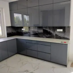 Nowoczesna kuchnia z szarymi, lakierowanymi frontami, blatem w kolorze jasnego betonu i dekoracyjnym panelem ściennym imitującym marmur z widocznymi gniazdkami elektrycznymi oraz białymi płytkami...