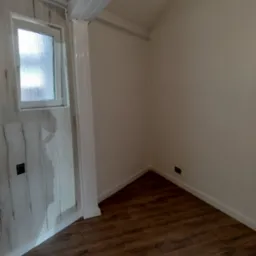 Wnętrze pokoju po remoncie: panele podłogowe imitujące drewno, ściany pomalowane na biało, okno z szybą ornamentową, proste oświetlenie sufitowe.