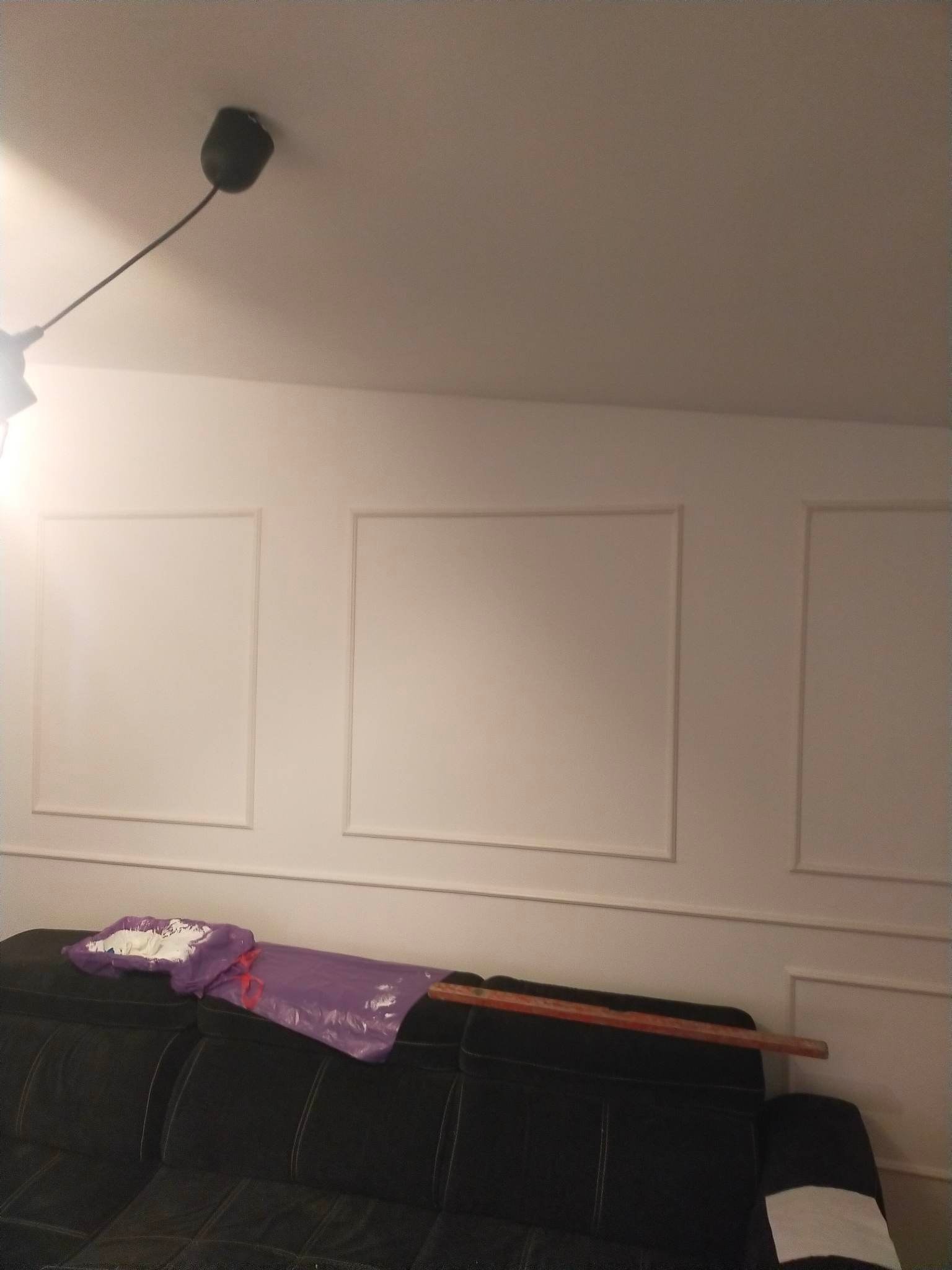 Biała ściana z dekoracyjnymi ramkami, czarna sofa z workiem farby i listwą przypodłogową, lampa sufitowa w rogu.